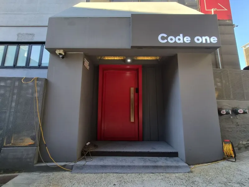 회색 캐노피 위 Code one 간판과 붉은 출입문이 있는 매장 입구 외관